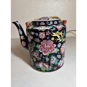 May include: A cylindrical ceramic teapot with a black base color, decorated with a vibrant floral pattern featuring pink, red, and green blossoms. It includes a curved spout, a matching lid, and a curved handle made of a light brown woven material.