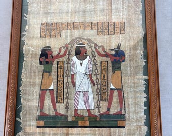 May include: Framed Egyptian papyrus art showing figures in traditional dress. The artwork features hieroglyphs and colourful illustrations of ancient Egyptian scenes. The frame is a dark brown colour.