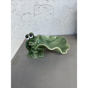 Può includere: Una statuetta in ceramica verde lucido raffigurante una rana che regge una grande foglia curva che forma una ciotola poco profonda. La rana ha occhi bianchi prominenti con pupille nere e la foglia presenta venature dettagliate in rilievo.