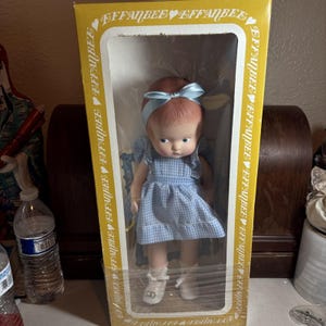 May include: A doll with reddish-brown hair and a light blue ribbon headband, wearing a light blue and white gingham dress with ruffled sleeves and white shoes. The doll is inside a yellow rectangular box with the text EFFANBEE printed repeatedly.