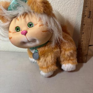 Peut inclure: Une poupée chat en peluche orange et blanche vintage avec des yeux verts, un nez rose et un nœud bleu sarcelle. La poupée a un collier vert avec une étiquette argentée. Le chat a des pattes blanches et une queue blanche.