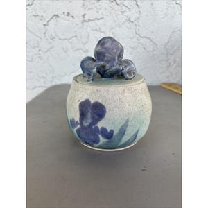 May include: A round ceramic lidded jar with a speckled off-white and light blue glaze. The lid features a sculpted three-dimensional purple iris flower as a handle. The body of the jar is decorated with a painted dark blue iris flower design.