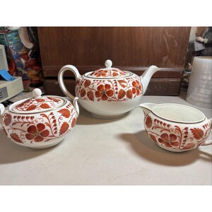 Pode incluir: Um conjunto de chá em cerâmica branca composto por um bule com tampa, um açucareiro com tampa e uma leiteira. Cada peça apresenta um padrão repetitivo de flores e folhas vermelho-alaranjadas com detalhes em dourado nas bordas.