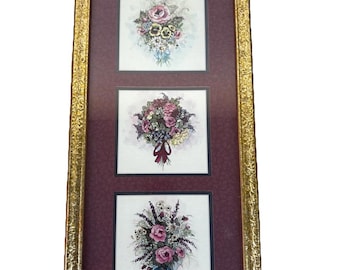 May include: A framed floral art piece featuring three square images of bouquets. The bouquets are composed of pink, purple, and white flowers with green foliage. The frame is gold with an ornate design, and the background is a deep burgundy.