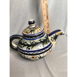 May include: A ceramic teapot with a lid, featuring hand-painted black, blue, and orange floral patterns on a cream background. A wooden ruler is positioned vertically behind the teapot, showing a height measurement of approximately 6 inches.