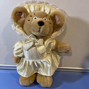 May include: A plush teddy bear wearing a cream-colored dress and bonnet. The bear has a brown face with black eyes and a black nose. The dress features a bow at the waist. The bonnet is trimmed with lace.
