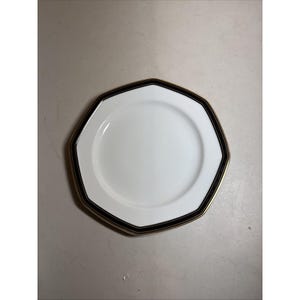 Op de afbeelding: Een achthoekig wit keramieken bord met een brede zwarte rand langs de zijkant, geaccentueerd door een dunne goudkleurige lijn aan de buitenste rand. Het midden van het bord is effen wit en de vorm wordt bepaald door acht zijden.