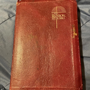 May include: A well-worn, red leather-bound book titled "The Rainbow Study Bible." The cover features the title in gold lettering with a stylized rainbow graphic. The book shows signs of age and use.