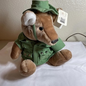 May include: A plush teddy bear wearing a green safari-style outfit, including a hat and jacket with pockets. The bear has brown fur, a black nose, and a white ear. A tag is attached to the bear.