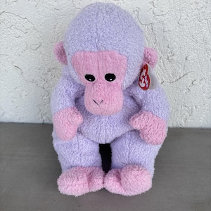 May include: A plush gorilla toy with lavender fur and light pink accents on the face, hands, and feet. The stuffed animal features black plastic eyes and a small red heart-shaped Ty tag attached to the side of its head near the ear.