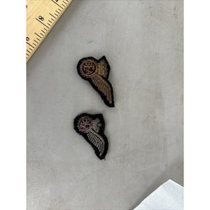 May include: Two embroidered patches featuring a central maroon circular emblem with a crown and wings extending to the side. The patches are black with gold-toned metallic thread detailing. A wooden ruler is visible, showing a measurement of 8 inches.