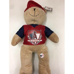 May include: A plush teddy bear wearing a red cap and a red and navy blue jersey with the Los Angeles Angels logo. The bear is tan with a stitched black mouth and a white MLB logo button.