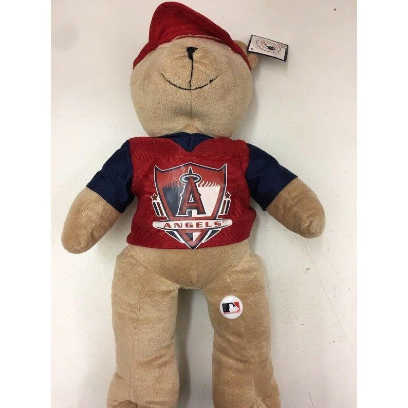 May include: A plush teddy bear wearing a red cap and a red and navy blue jersey with the Los Angeles Angels logo. The bear is tan with a stitched black mouth and a white MLB logo button.