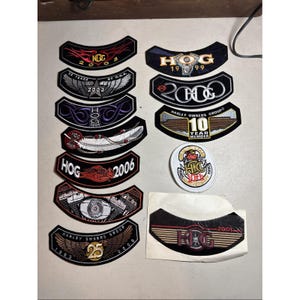 May include: A collection of Harley Owners Group (HOG) patches in various shapes and designs. The patches feature text such as "HOG," "2006," and "25 Years." Colors include black, red, white, and gold. Some patches have wings or motorcycle engine graphics.
