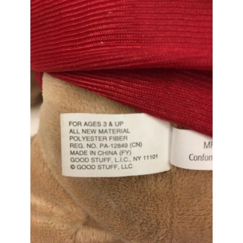 May include: Close-up of a plush toy with a red fabric and a tan fabric. A white tag reads "FOR AGES 3 & UP", "ALL NEW MATERIAL", "POLYESTER FIBER", and "MADE IN CHINA (FY)".