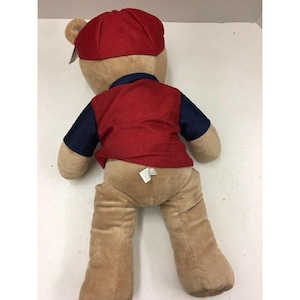 May include: A plush teddy bear wearing a red baseball cap, a red vest, and a navy blue shirt. The bear is light brown and has a tag attached to its ear. The bear is lying down.