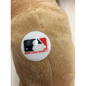 May include: A close-up of a plush, tan-colored object with a white, circular button attached. The button features the Major League Baseball logo in black, red, and blue, with the text "MAJOR LEAGUE BASEBALL" in a red font.