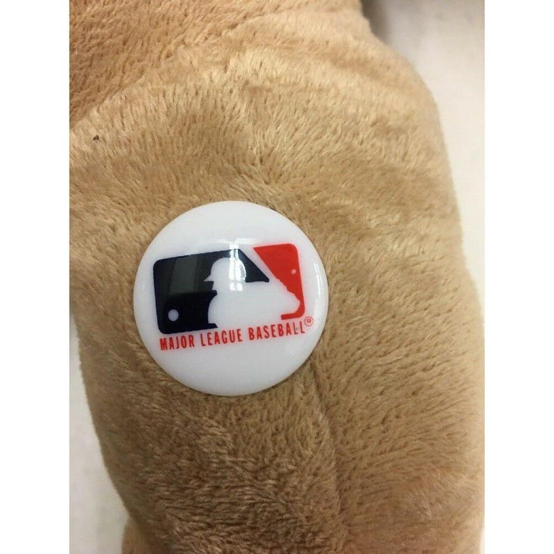 May include: A close-up of a plush, tan-colored object with a white, circular button attached. The button features the Major League Baseball logo in black, red, and blue, with the text "MAJOR LEAGUE BASEBALL" in a red font.