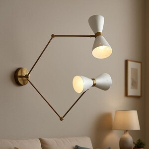 May include: A contemporary wall light with two white, hourglass-shaped shades and brass-coloured arms. The light is mounted on a neutral-coloured wall above a beige sofa with decorative cushions. A table lamp and framed artwork are also visible.