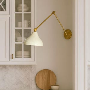 May include: A kitchen interior with a cream-coloured, adjustable wall-mounted lamp with a brass finish. The lamp is above a countertop with a wooden chopping board and a small bowl. White cabinets and a marble backsplash are also visible.