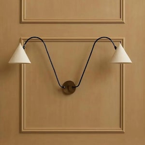 Può includere: Applique da parete moderno con due paralumi conici. I paralumi sono bianchi e l'apparecchio ha un design a braccio nero ondulato. Al centro c'è una piastra circolare in ottone. L'applique è montata su una parete beige con modanature decorative.