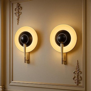 Puede incluir: Dos apliques de pared con un diseño circular en blanco y negro, con un anillo de luz crema y una base dorada. Los apliques tienen una estética moderna de mediados de siglo, montados en una pared beige con molduras doradas decorativas.
