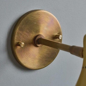 May include: Close-up of a brass wall sconce. The sconce features a circular backplate with two small screws and a central rod extending to the light fixture. The brass has a warm, aged finish, suggesting a vintage or antique style. The fixture is mounted on a light gray wall.