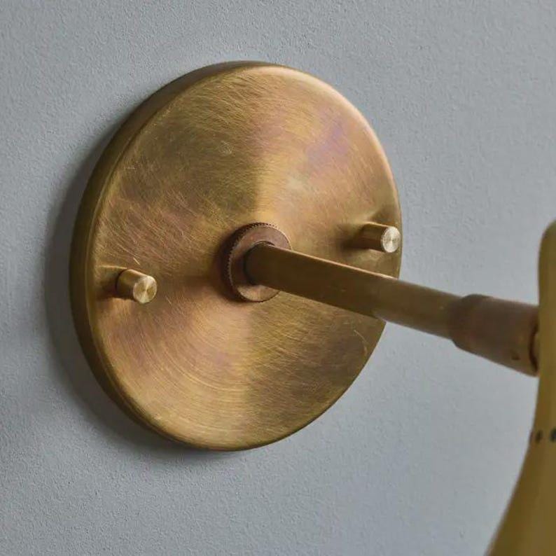 May include: Close-up of a brass wall sconce. The sconce features a circular backplate with two small screws and a central rod extending to the light fixture. The brass has a warm, aged finish, suggesting a vintage or antique style. The fixture is mounted on a light gray wall.
