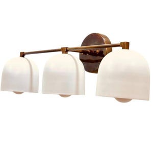 May include: A three-light vanity fixture with a bronze finish. It features three white, bell-shaped shades and a round backplate. The fixture is designed for bathroom or vanity lighting.