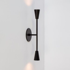 May include: A black wall sconce with two conical light holders. The sconce is mounted on a white wall and has a minimalist design. The light fixtures are positioned vertically, creating a sleek, contemporary look. The sconce is made of metal.