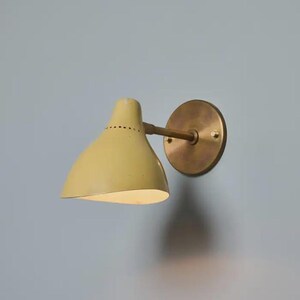 May include: A wall-mounted light fixture with a pale yellow conical shade and a brass-colored circular backplate. The shade has a series of small holes near the top. The fixture is mounted on a light blue wall.