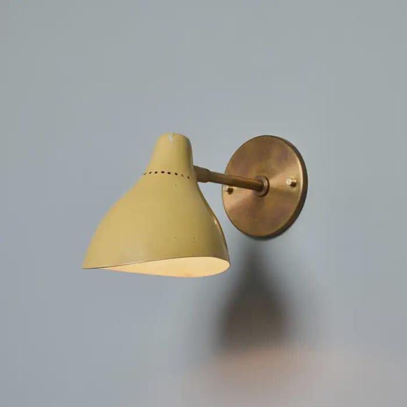 May include: A wall-mounted light fixture with a pale yellow conical shade and a brass-colored circular backplate. The shade has a series of small holes near the top. The fixture is mounted on a light blue wall.