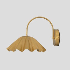 May include: A gold wall sconce featuring a pleated, scalloped shade and a curved arm. The fixture is mounted on a circular base. The metal sconce has a warm gold finish, adding a touch of elegance to any interior space. The shade is approximately 30 cm in diameter.