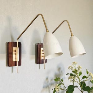 Peut inclure: Deux appliques murales de style moderne du milieu du siècle. Elles présentent une base en bois foncé, des détails en laiton et des abat-jours blancs en forme de cône. Les appliques sont fixées sur un mur clair.