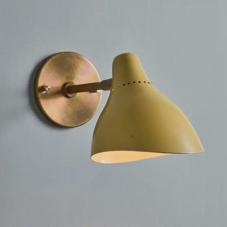 May include: A wall-mounted light fixture with a mustard yellow shade and a brass-colored mounting plate and arm. The shade has a curved, conical shape with small perforations near the top. The fixture is designed for indoor use.