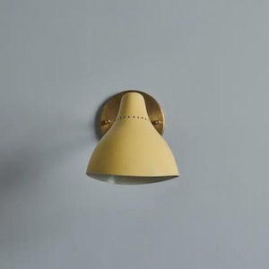 May include: A yellow wall sconce with a brass-colored mounting plate. The sconce has a conical shade with a series of small holes near the top. The fixture is mounted on a light blue wall.