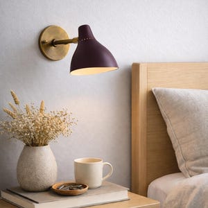 May include: A wall-mounted lamp with a burgundy shade and brass accents illuminates a bedside table. A vase of dried flowers, a mug, and a small wooden dish sit on the table next to a book. The scene is set in a bedroom with a wooden bed frame.