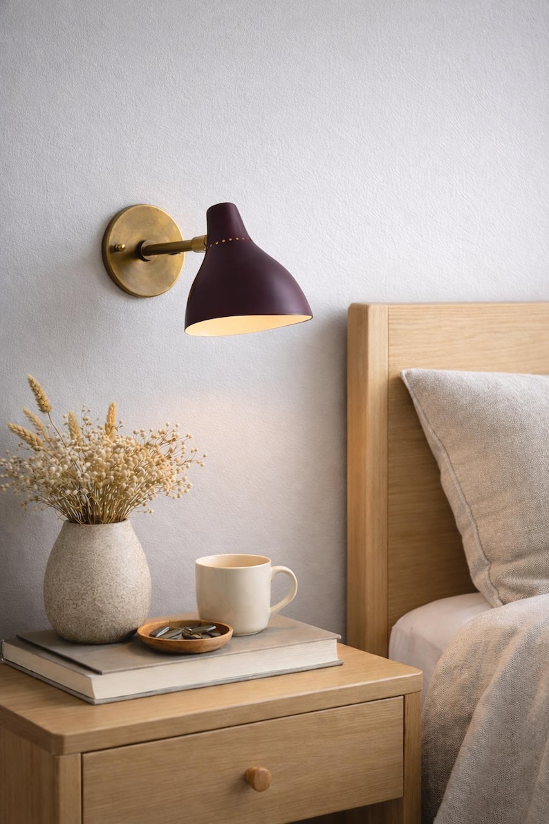 May include: A wall-mounted lamp with a burgundy shade and brass accents illuminates a bedside table. A vase of dried flowers, a mug, and a small wooden dish sit on the table next to a book. The scene is set in a bedroom with a wooden bed frame.