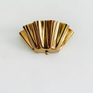 May include: A gold-colored, fan-shaped wall sconce with a small, round detail at the bottom. The sconce has a series of vertical, curved ridges, creating a textured, decorative design. The sconce is mounted on a white wall.