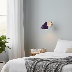 May include: A modern wall-mounted lamp with a deep purple shade and a brass-colored circular base. The lamp is positioned above a bed with white linens and a gray knitted throw. A potted plant is visible in the background.
