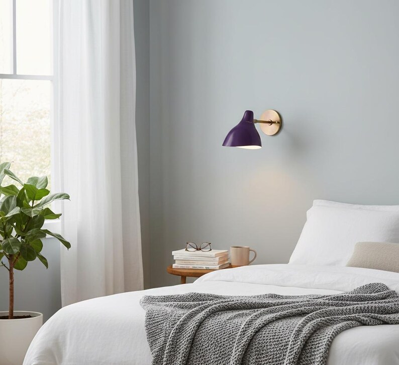 May include: A modern wall-mounted lamp with a deep purple shade and a brass-colored circular base. The lamp is positioned above a bed with white linens and a gray knitted throw. A potted plant is visible in the background.