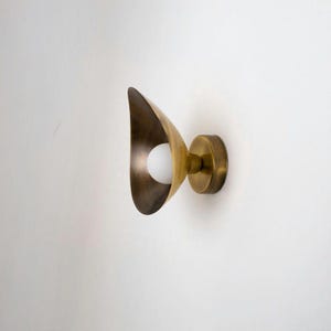 May include: A brass wall sconce with a unique, curved design. The fixture features a rounded base and a flared, leaf-like shade that houses a white, spherical light bulb. The sconce is mounted on a white wall, showcasing its warm metallic finish.