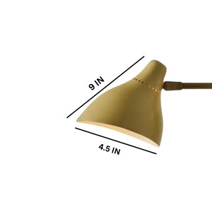 May include: A wall-mounted lamp with a pale yellow shade, measuring 9 inches long and 4.5 inches wide. The lamp has a brass arm and a circular mounting plate. The shade has small perforations along the top edge.