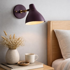 May include: A plum-colored wall-mounted lamp with a brass arm and a round base. The lamp is positioned above a wooden nightstand with a vase of dried flowers, a mug, and a small wooden dish. A bed with a wooden headboard is in the background.