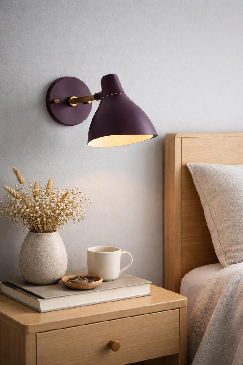 May include: A plum-colored wall-mounted lamp with a brass arm and a round base. The lamp is positioned above a wooden nightstand with a vase of dried flowers, a mug, and a small wooden dish. A bed with a wooden headboard is in the background.