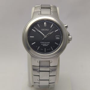 May include: A silver Seiko Presage Kinetic 100M watch with a black dial. The watch features a date display at the 3 o'clock position and silver hands and markers. The watch band is also silver.