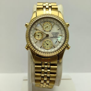 Könnte beinhalten: Goldfarbene Citizen Quartz Alarm Chronograph Uhr. Das weiße Zifferblatt hat mehrere Hilfszifferblätter, eine Datumsanzeige und goldene Zeiger und Markierungen. Sie hat ein goldenes Armband und eine strukturierte Lünette.
