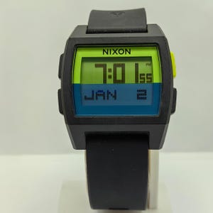 May include: Black digital watch with a rectangular face. The watch face displays the time as 7:01 PM and the date as Jan 2. The watch has a black band and the brand name "NIXON" is visible on the face.
