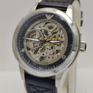Puede incluir: Reloj Emporio Armani plateado con correa de cuero negro. La esfera del reloj presenta un diseño esqueleto, mostrando los mecanismos internos, con las palabras "Emporio Armani" y "Meccanico". El reloj tiene una esfera redonda con marcadores de hora y minuto.