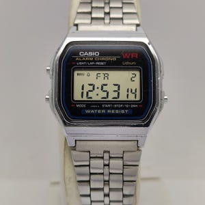 May include: A silver Casio digital watch with a black and blue face showing the time 12:53:14. The watch has a rectangular face with the Casio logo and the words "ALARM CHRONO" and "WATER RESIST". The watch band is made of silver metal.